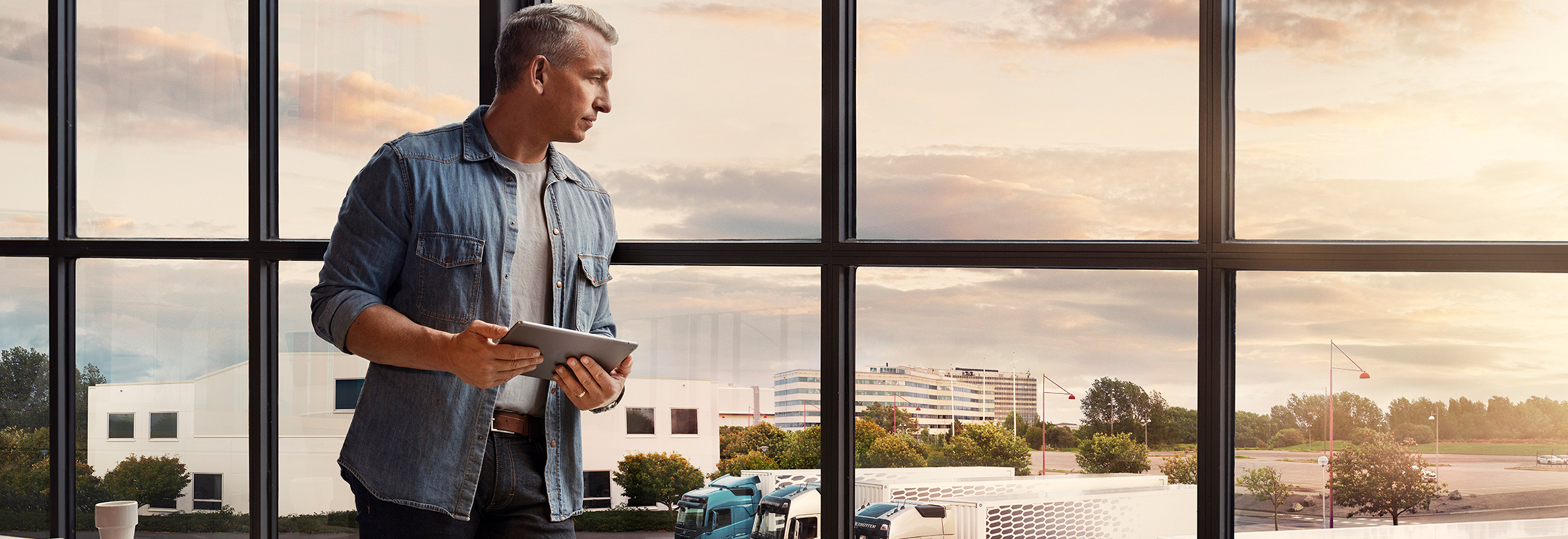 Un hombre que sostiene una tablet junto a una ventana y observa su flota de camiones